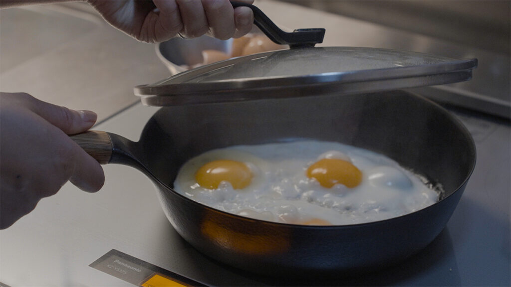 VERMICULARの24cm深型フライパンで目玉焼きを焼きながら、hふたを少し開けて焼き加減を確認する様子。蒸気の奥に見える黄身がほどよく火が通り、丁寧な調理の一場面。