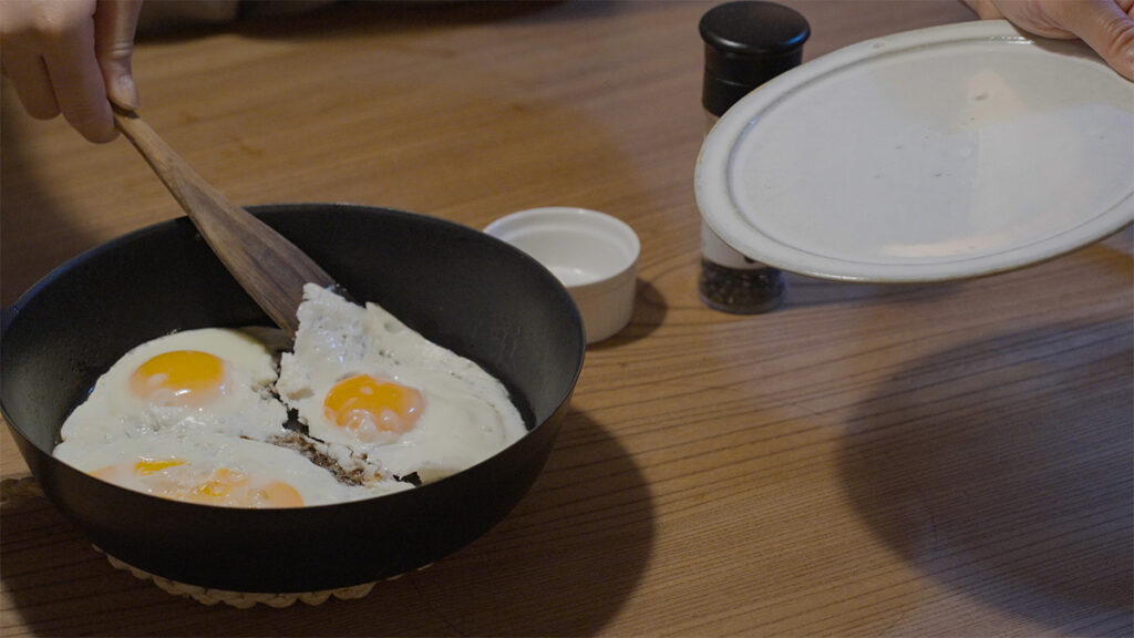 卓上に置かれたVERMICULARの24cm深型フライパンで焼いた目玉焼きを、木べらで皿に取り分けようとする様子。ふっくら焼き上がった卵をやさしくすくい上げる、食卓の穏やかな一場面。