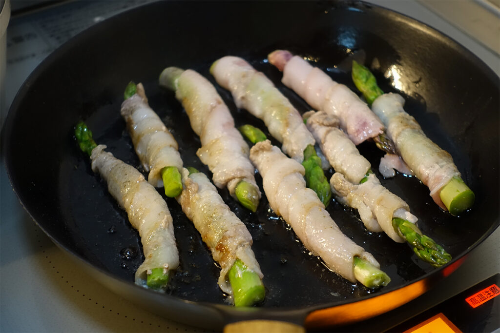 VERMICULARフライパン26サイズ浅型でアスパラの豚肉巻きを焼いている様子。表面がじゅわっと焼けて、肉の香ばしい香りが立ちのぼる。