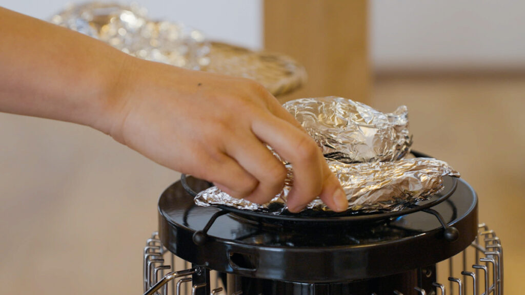 点火したストーブの上にアルミホイルで包んださつまいもをのせ、焼き芋の準備をしている様子を横から撮影した写真