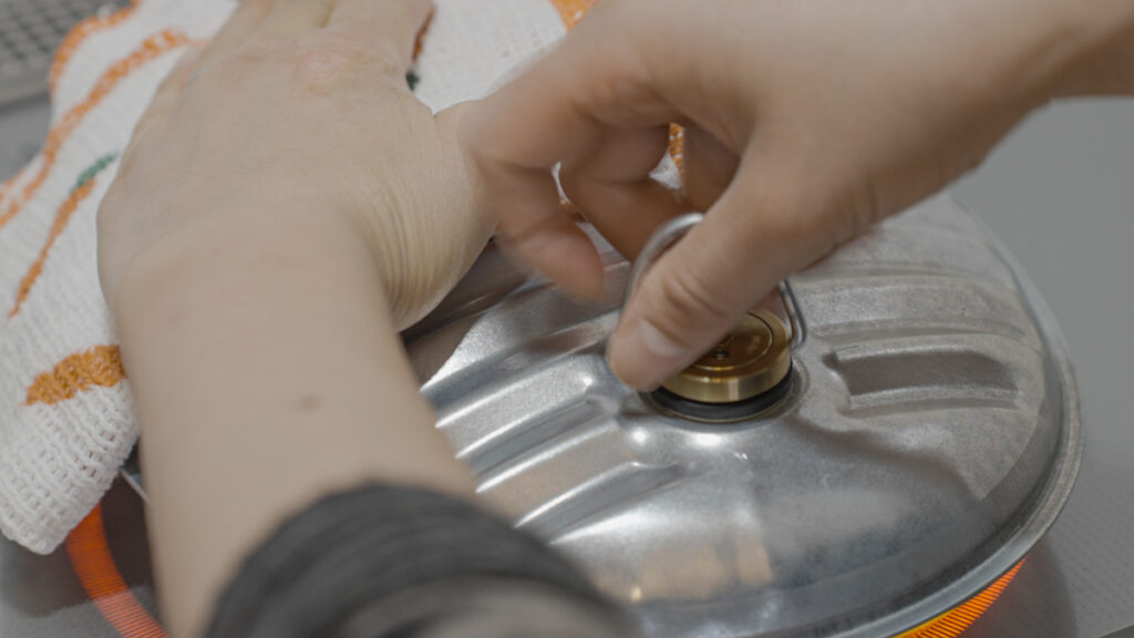 IHで温めたマルカの金属製湯たんぽに、やけどに注意しながら布巾で手を保護しつつ蓋を閉めている様子。金属製ならではの光沢と手元の動作がわかるシーン
